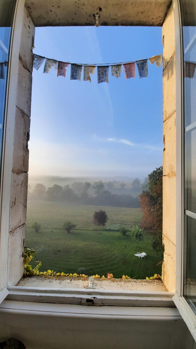 Une grande fenêtre ouverte en surplomb sur le paysage des causses du Quercy dans le brouillard. Drapeaux de prière en banderole en haut de la fenêtre.